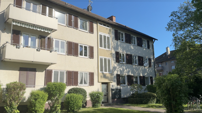 Residential apartment building with beige exterior, brown shutters, and multiple windows under a blue sky, with shrubs in front.