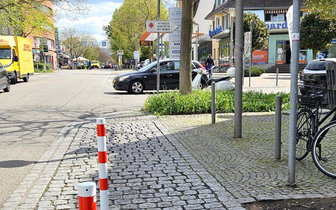 Street view in a European town: cobblestone sidewalk with red-white bollards, parked cars, storefronts, and a blue taxi sign.