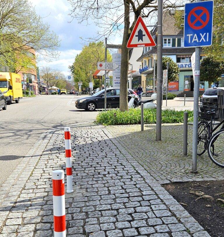 Street view in a European town: cobblestone sidewalk with red-white bollards, parked cars, storefronts, and a blue taxi sign.