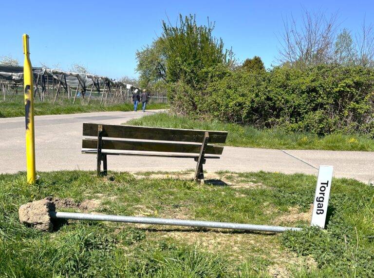 Two people walk along a paved path beside a hedge on a sunny day, with construction debris and a yellow post in the foreground.