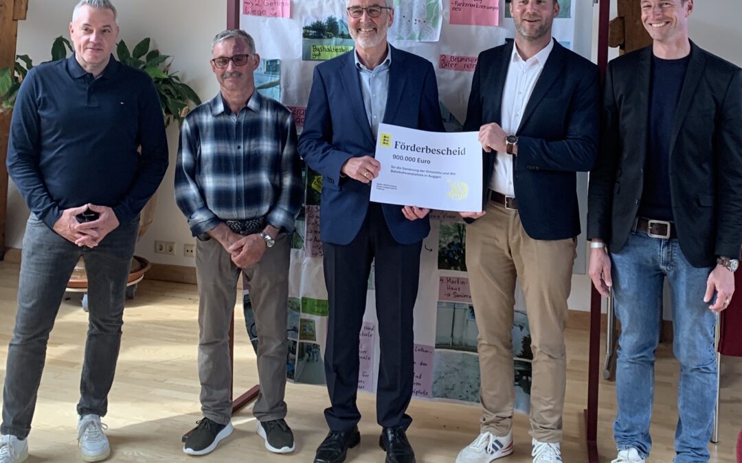 Five men stand indoors, the central man in a suit holds a large grant check while others smile behind him.