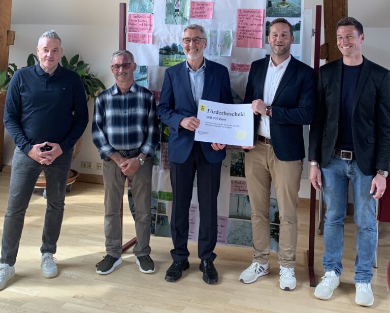 Five men stand indoors, the central man in a suit holds a large grant check while others smile behind him.