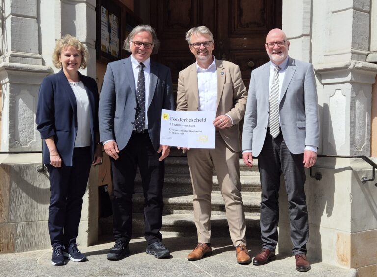 Four professionally dressed people stand on stone steps outside a building, holding a large Förderbescheid grant cheque.