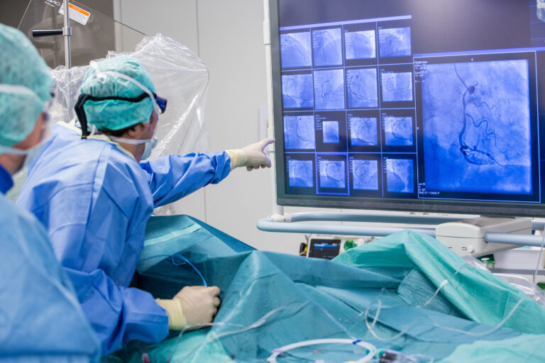 Surgeons in blue scrubs and masks monitor angiography images on a large screen during a procedure