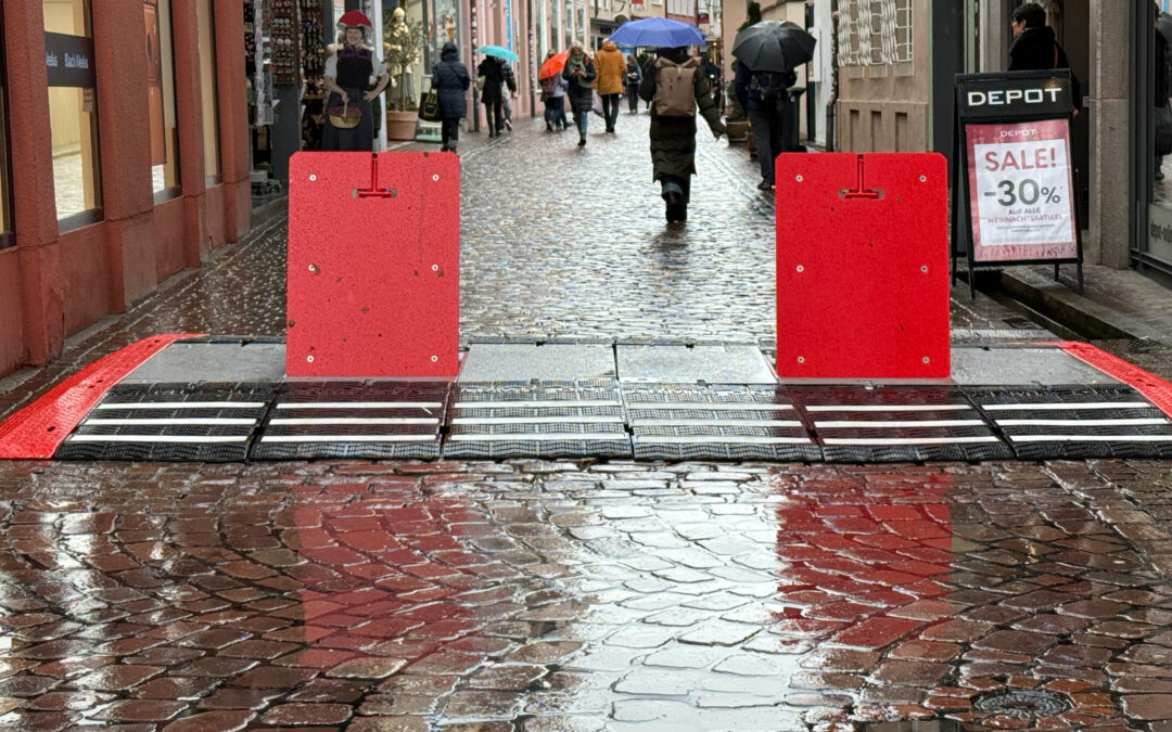 Pedestrians with umbrellas walking down a wet, cobblestone street blocked by red barriers in a narrow shopping lane.