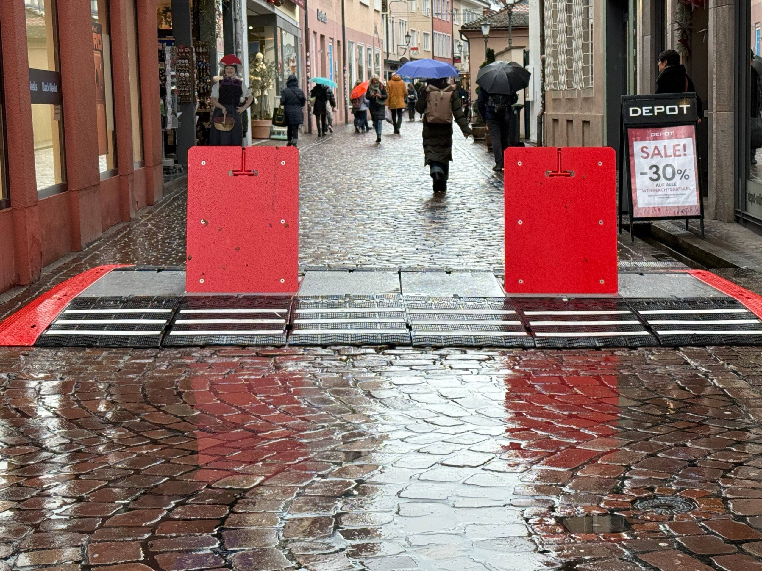 Pedestrians with umbrellas walking down a wet, cobblestone street blocked by red barriers in a narrow shopping lane.