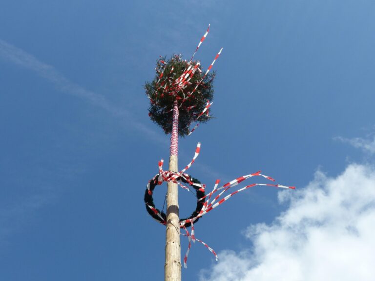 Tall wooden maypole adorned with greenery and red-and-white ribbons fluttering in the wind against a clear blue sky