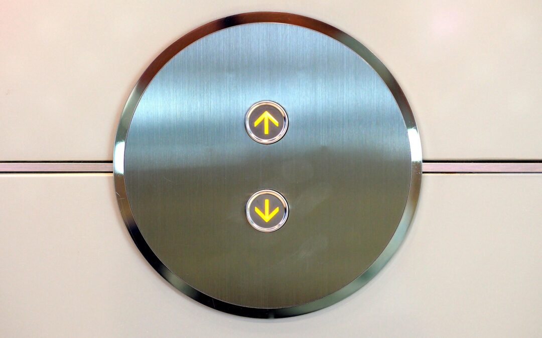 Circular brushed-metal elevator control panel with a lit yellow up-arrow button central on the circle (and a down-arrow below).