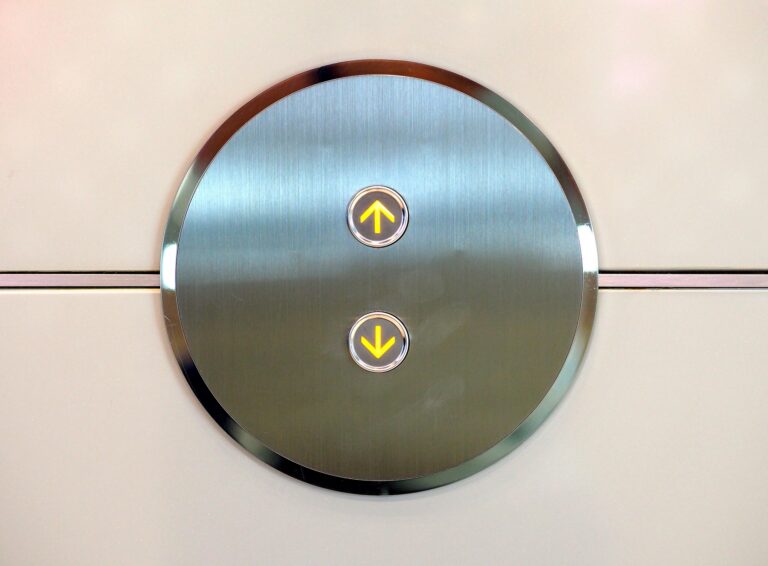 Circular brushed-metal elevator control panel with a lit yellow up-arrow button central on the circle (and a down-arrow below).