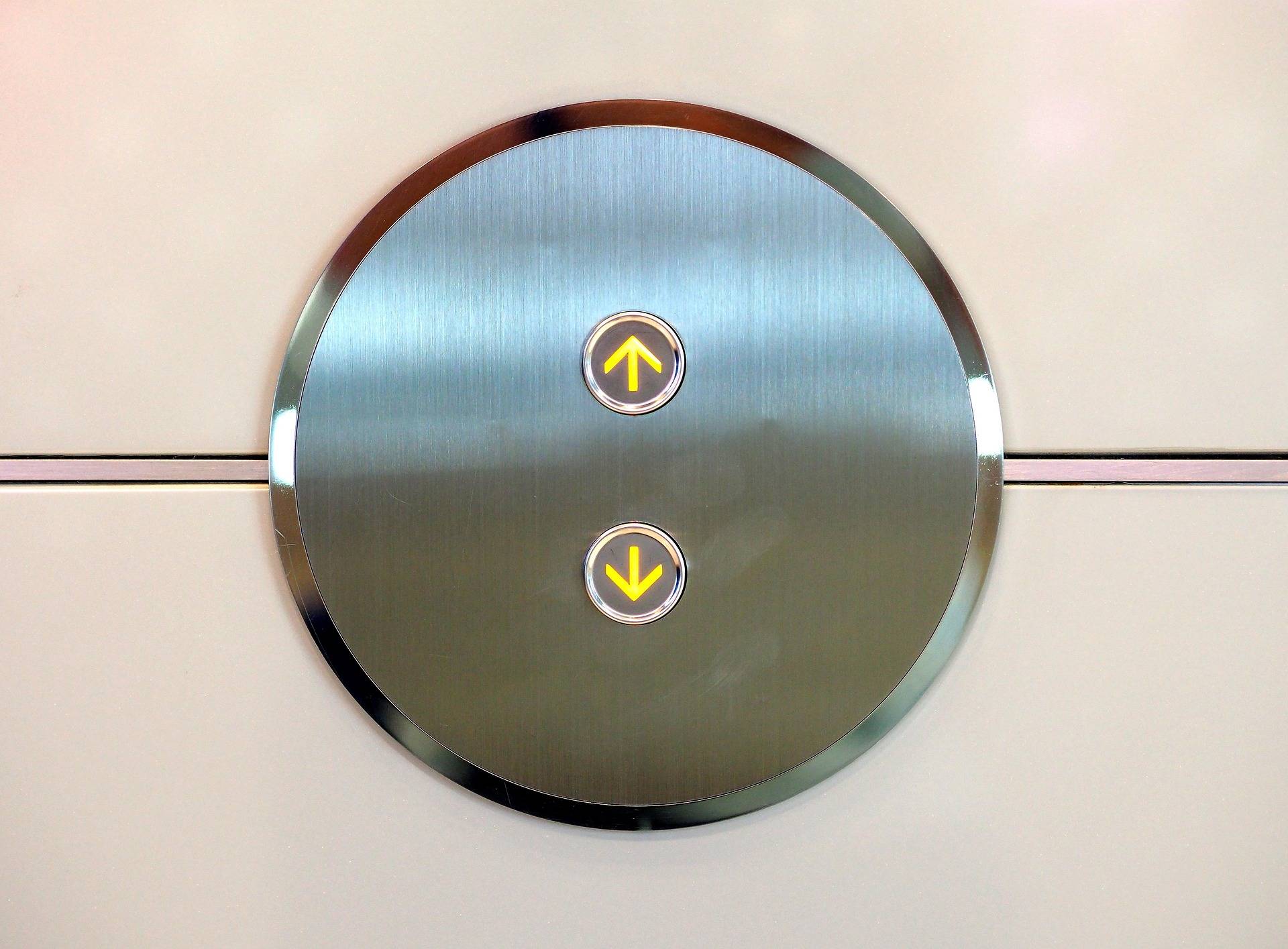 Circular brushed-metal elevator control panel with a lit yellow up-arrow button central on the circle (and a down-arrow below).