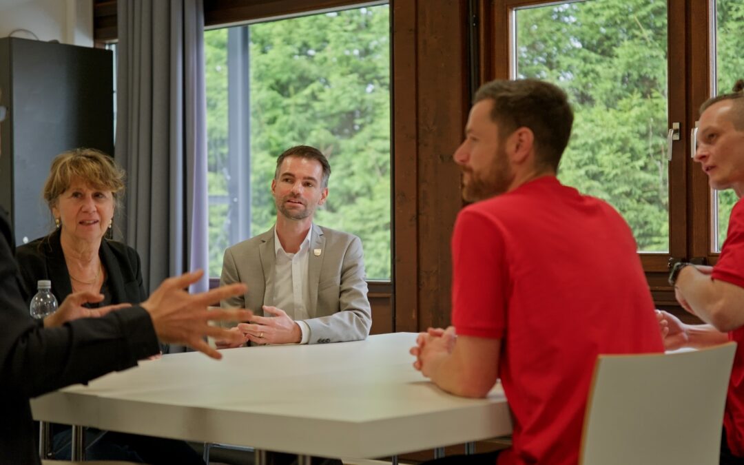 Five adults seated around a white table discussing in a bright room with large windows and greenery outside.
