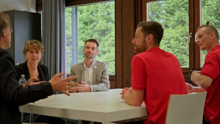 Five adults seated around a white table discussing in a bright room with large windows and greenery outside.