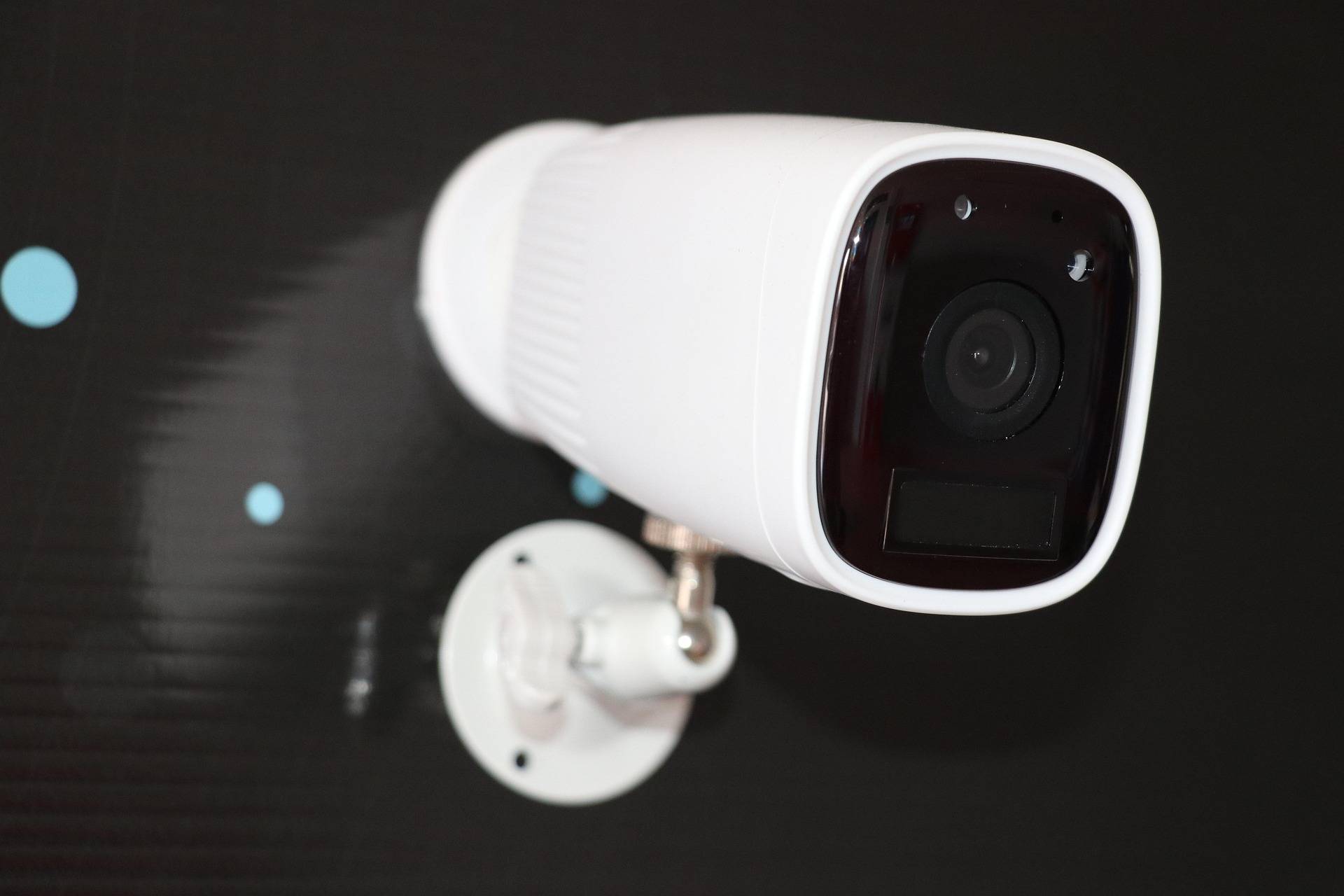 White dome security camera mounted on a wall with a dark lens panel facing forward, against a black background.
