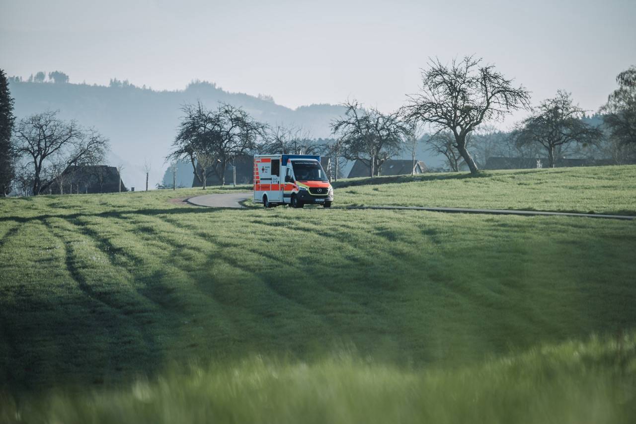 Orange and white ambulance parked on a curved road through a grassy rural landscape with leafless trees in the background.