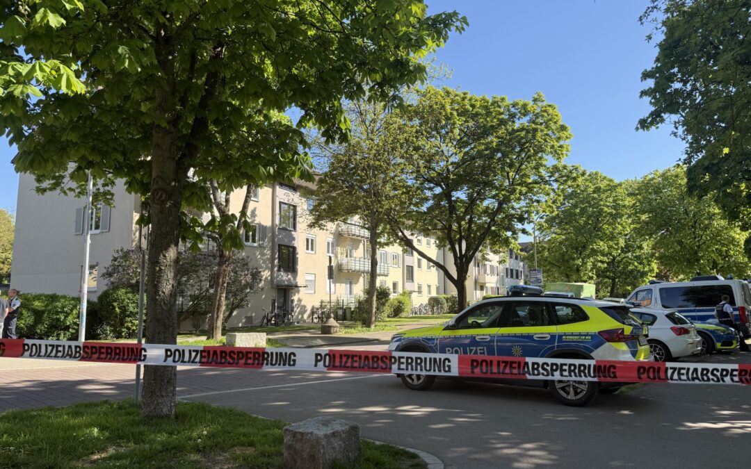 Police barrier tape blocks a residential street; police cars parked by a beige apartment building under leafy trees on a sunny day.
