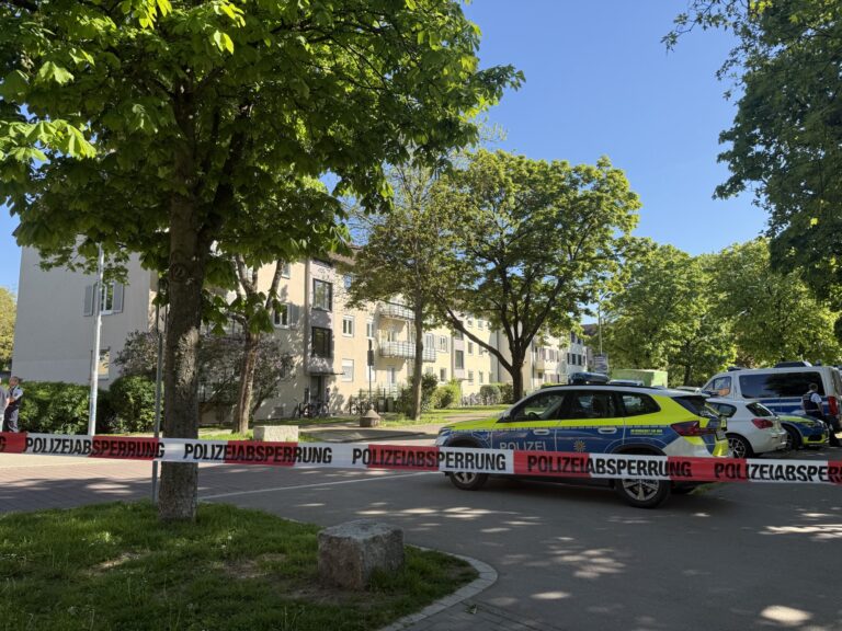 Police barrier tape blocks a residential street; police cars parked by a beige apartment building under leafy trees on a sunny day.