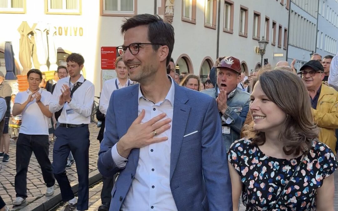 Smiling man in a blue suit places a hand on his chest while walking beside a woman in a patterned dress, as a crowd applauds behind them on a cobblestone street.