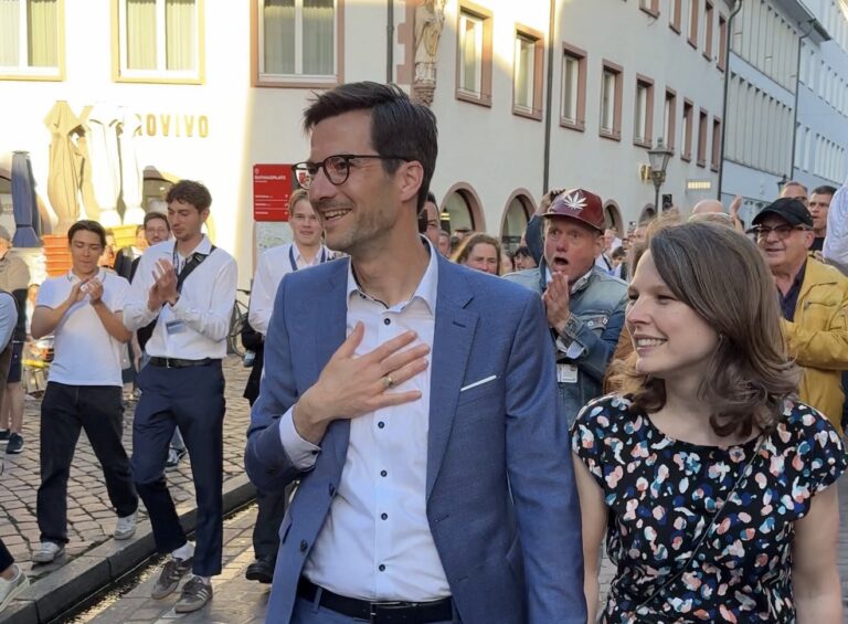 Smiling man in a blue suit places a hand on his chest while walking beside a woman in a patterned dress, as a crowd applauds behind them on a cobblestone street.