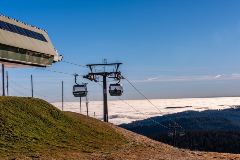 Cable car gondola lifts travel along a steel cable over a grassy hillside with a large angular station to the left and a clear blue sky above.