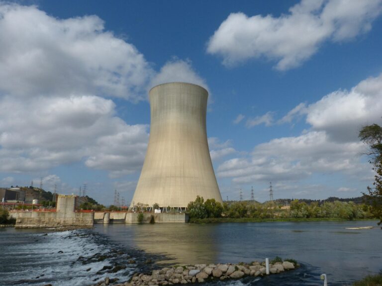 Large concrete cooling tower rising above a river, with rocky shoreline and distant industrial buildings under a blue, partly cloudy sky.
