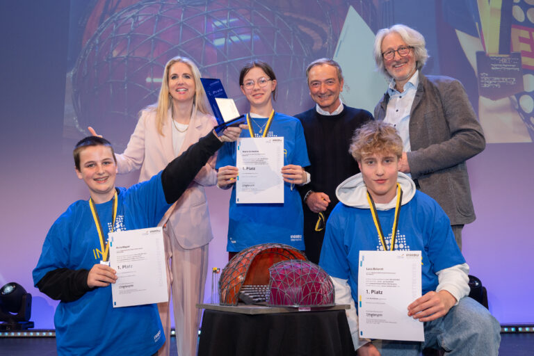 Group of young award recipients on stage posing with certificates and medals, a woman and man flanking them smile warmly as a glass trophy sits nearby on a table behind them.
