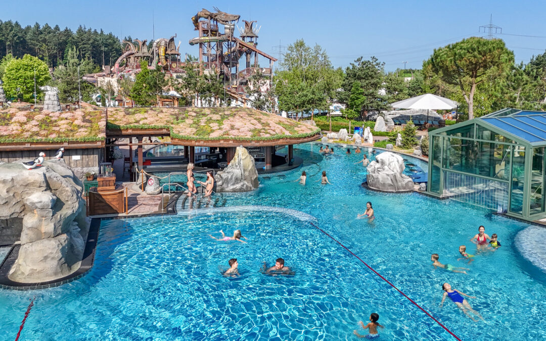 A large outdoor water park pool with slide towers, rocks, and people swimming on a sunny day.