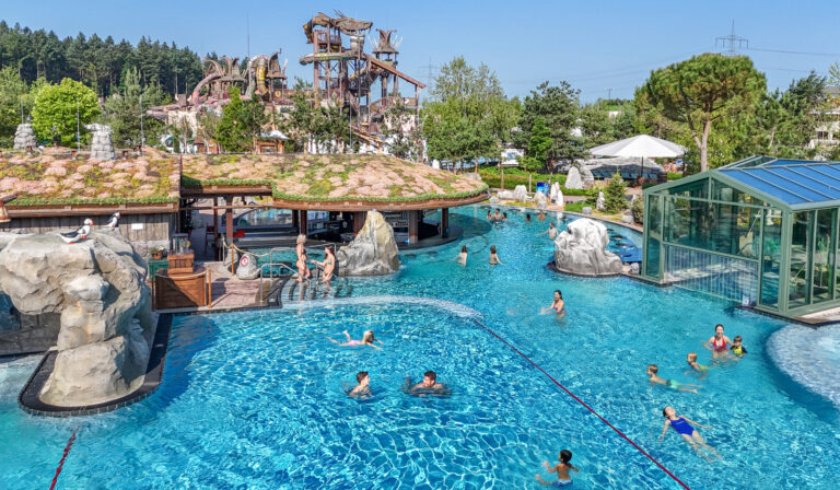 A large outdoor water park pool with slide towers, rocks, and people swimming on a sunny day.