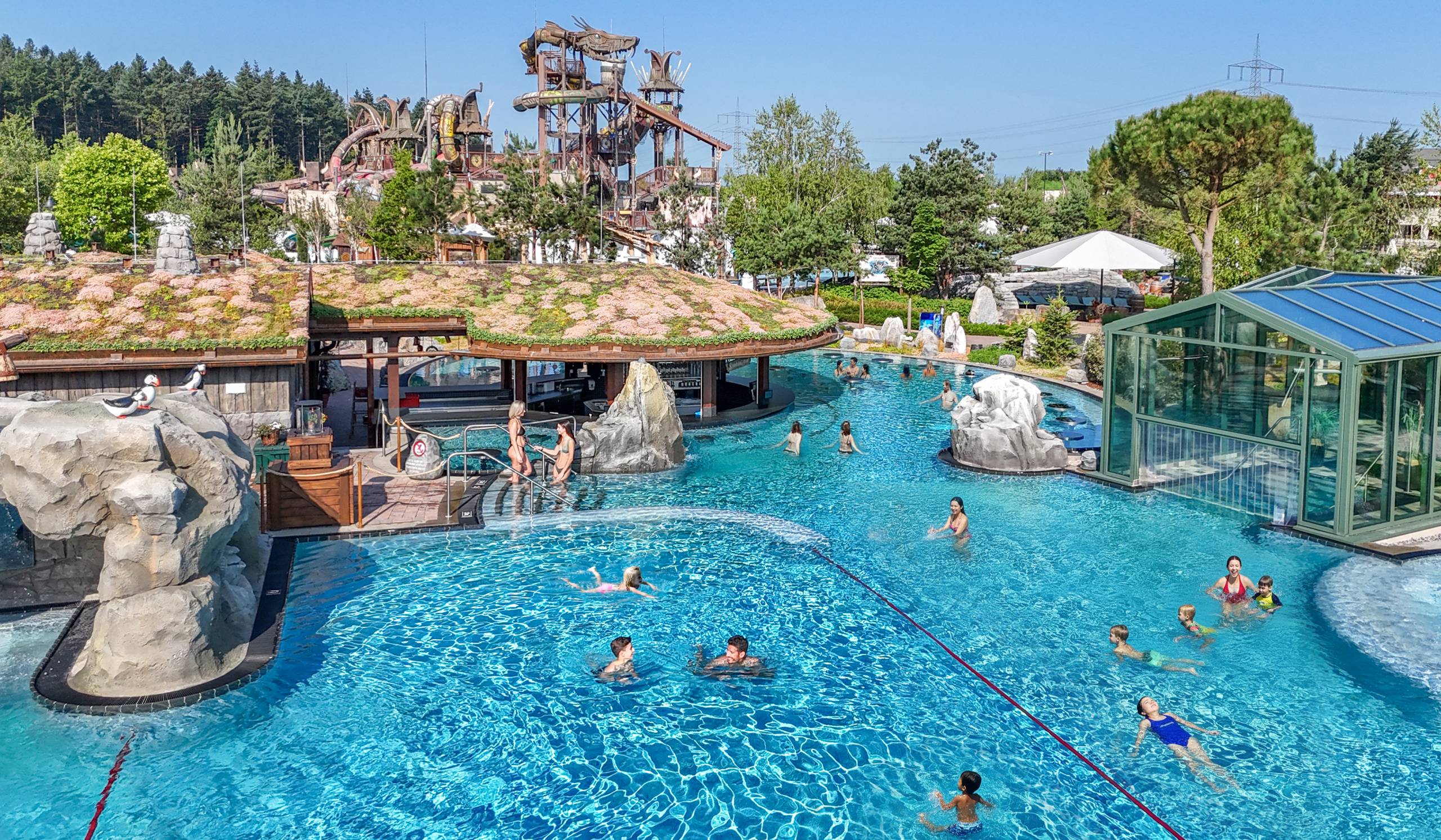 A large outdoor water park pool with slide towers, rocks, and people swimming on a sunny day.