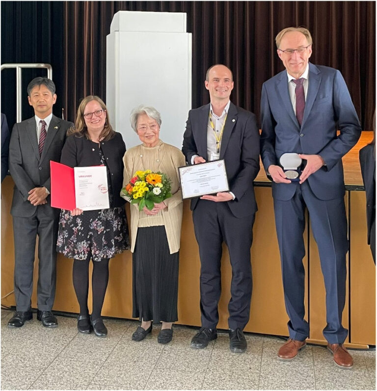 Five people in formal attire pose on a stage; a woman holds a red certificate folder, another holds a bouquet, and two men hold certificates/medal boxes.