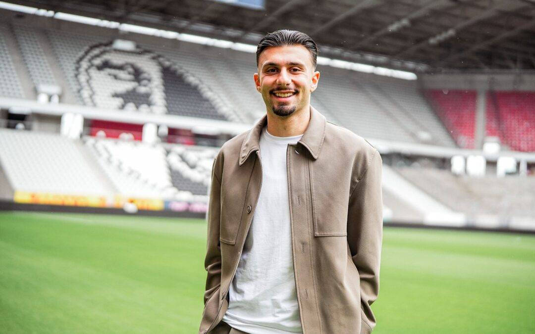 Smiling man in a beige jacket and white shirt stands on a green stadium field with blurred seating behind him.