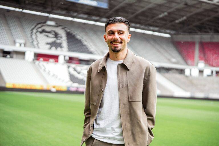 Smiling man in a beige jacket and white shirt stands on a green stadium field with blurred seating behind him.