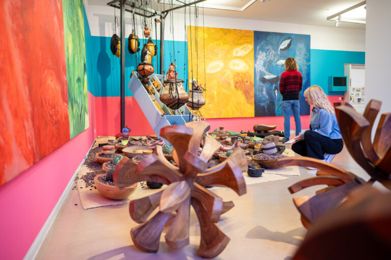 Gallery interior featuring vivid wall murals in pink, blue, and yellow with large curved wooden sculptures in the foreground and visitors viewing the exhibits.