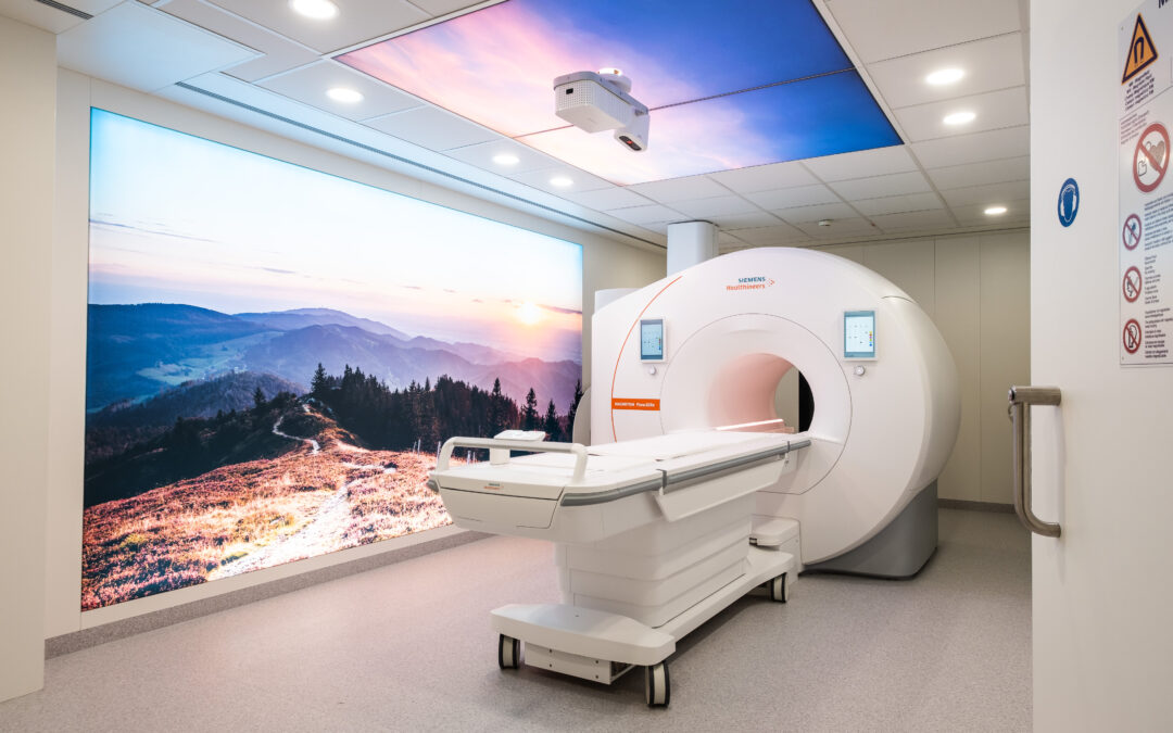 MRI scanner in a bright clinical room with a large wall mural of a mountain landscape at sunset behind it, depicting a medical imaging suite