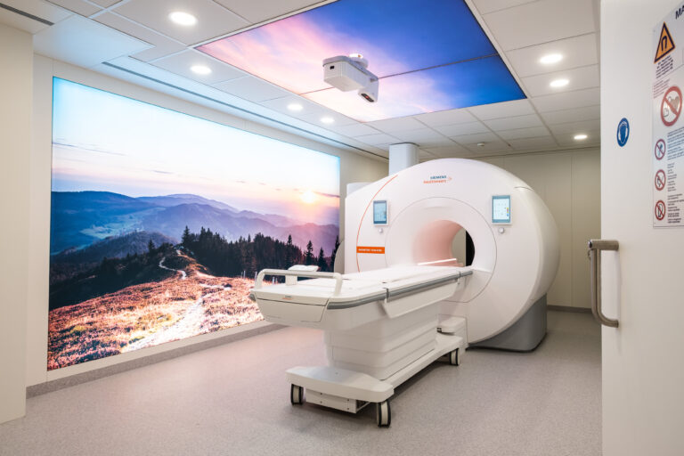 MRI scanner in a bright clinical room with a large wall mural of a mountain landscape at sunset behind it, depicting a medical imaging suite