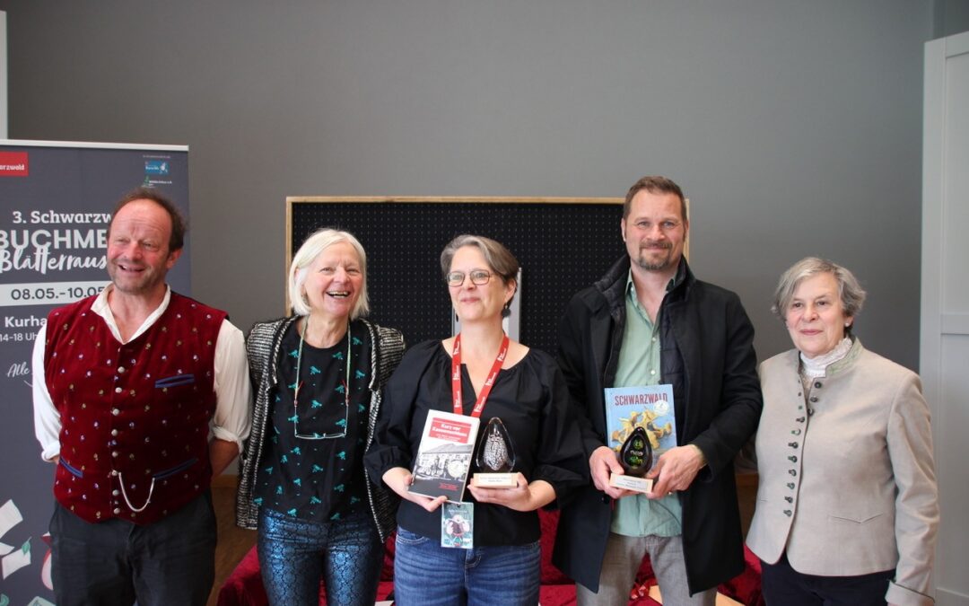 Group of five adults posing indoors, two holding books and a trophy, smiling after an awards event.