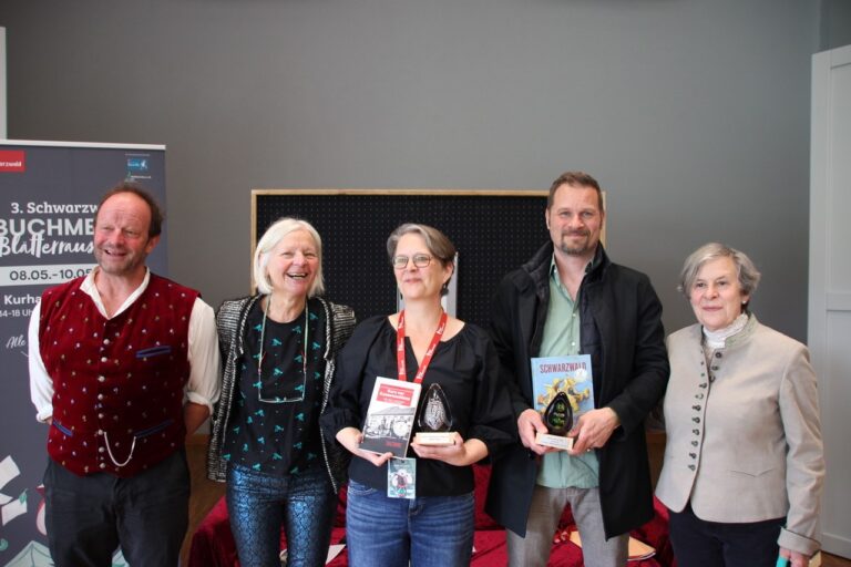 Group of five adults posing indoors, two holding books and a trophy, smiling after an awards event.