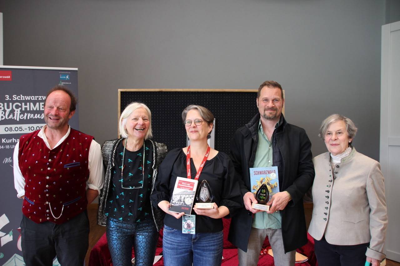 Group of five adults posing indoors, two holding books and a trophy, smiling after an awards event.