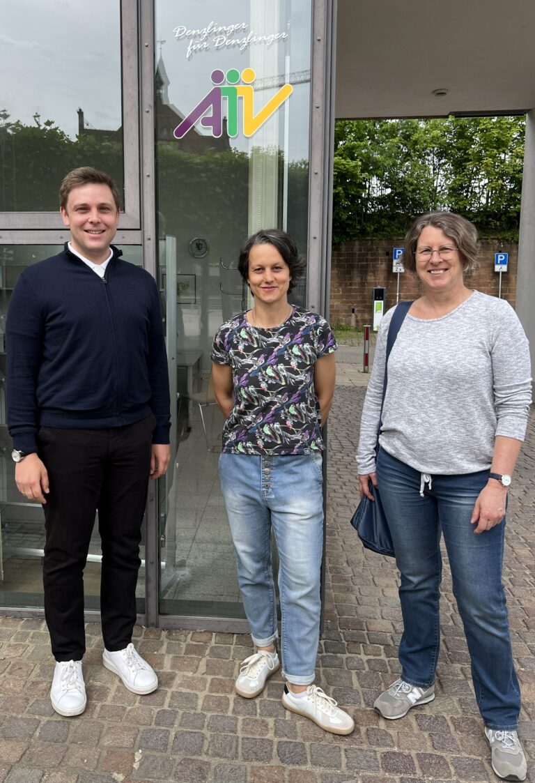 Three adults stand outside a glass building entrance with a colorful 'AIV' logo above them. Everyone smiles casually, wearing casual clothes and sneakers.