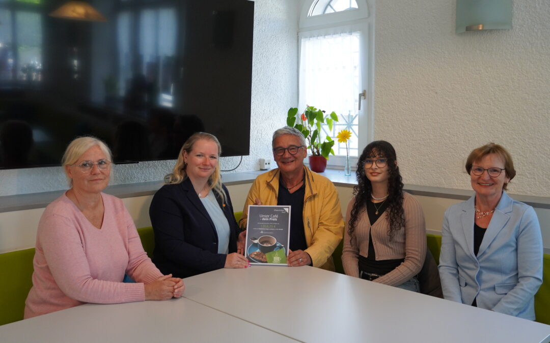 Group of five adults seated around a white table in a bright room, two women on left, a smiling man in a yellow jacket holding a brochure toward the camera with two younger women and another woman on the right side.