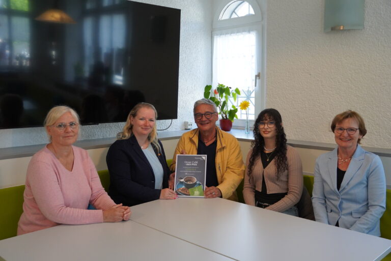 Group of five adults seated around a white table in a bright room, two women on left, a smiling man in a yellow jacket holding a brochure toward the camera with two younger women and another woman on the right side.