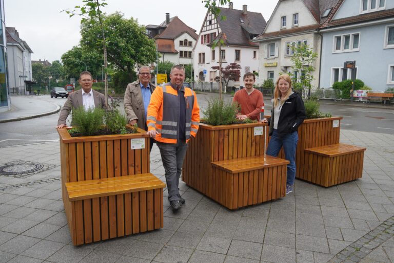Five people posing behind tall wooden planter boxes with greenery along a city street; one man wears an orange safety vest at center.