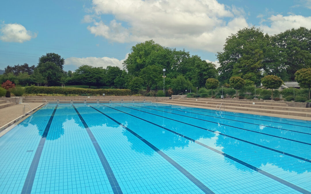 Outdoor swimming pool with blue tiled lanes and empty deck, surrounded by trees under a partly cloudy sky.