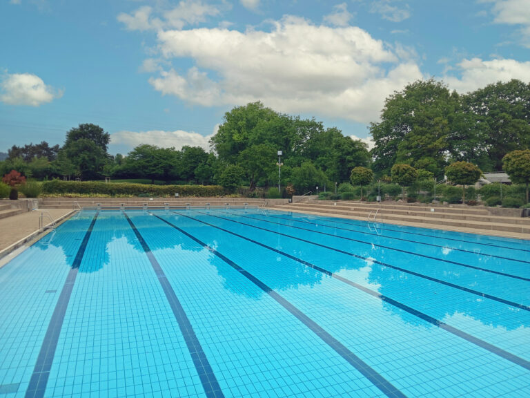 Outdoor swimming pool with blue tiled lanes and empty deck, surrounded by trees under a partly cloudy sky.