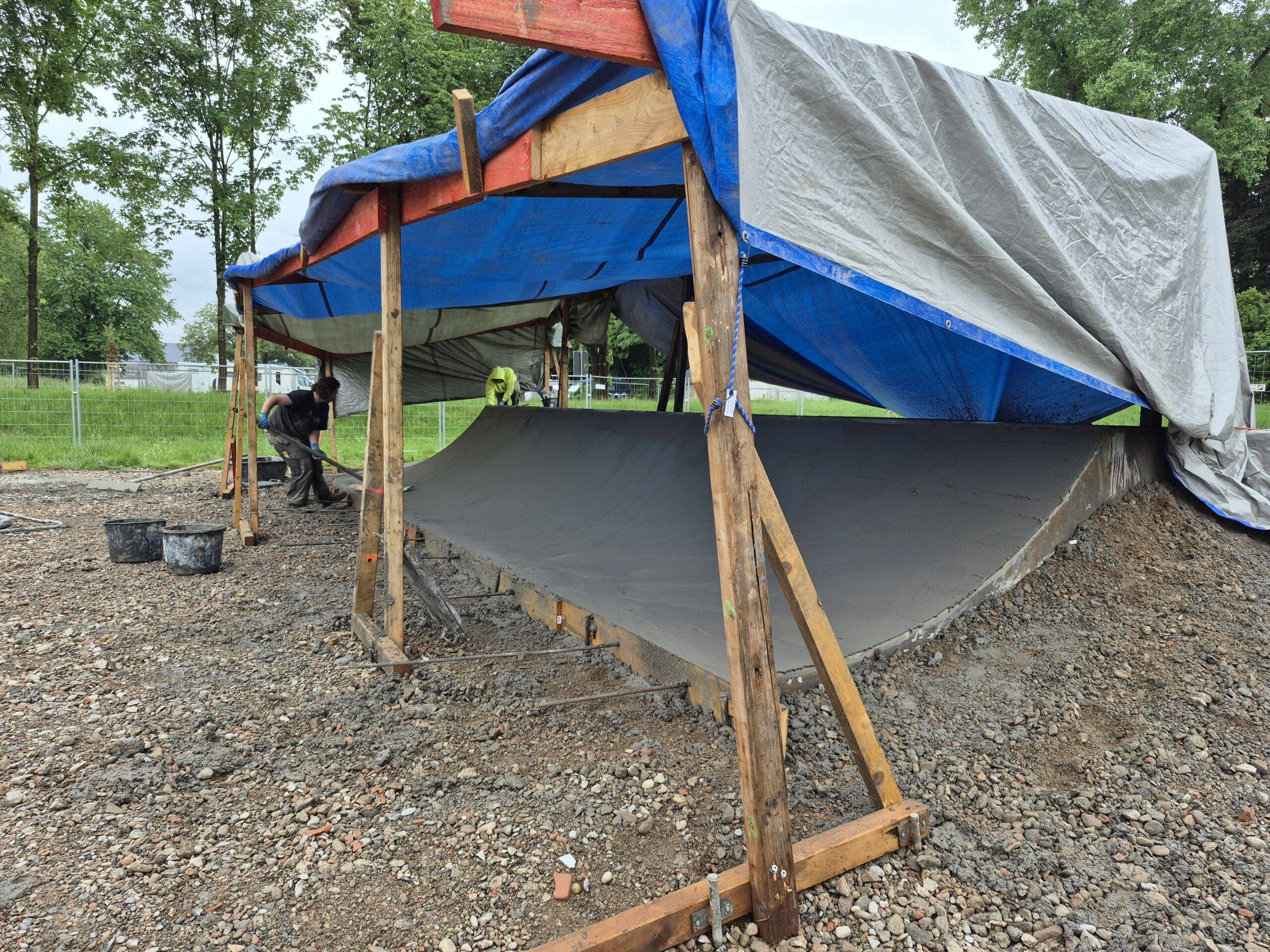 Construction site with wooden braces and a curved concrete form under blue and gray tarps.