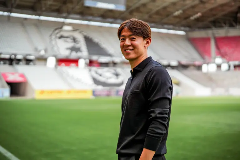 Smiling man in a dark polo standing on a green stadium field with blurred empty stands in the background.