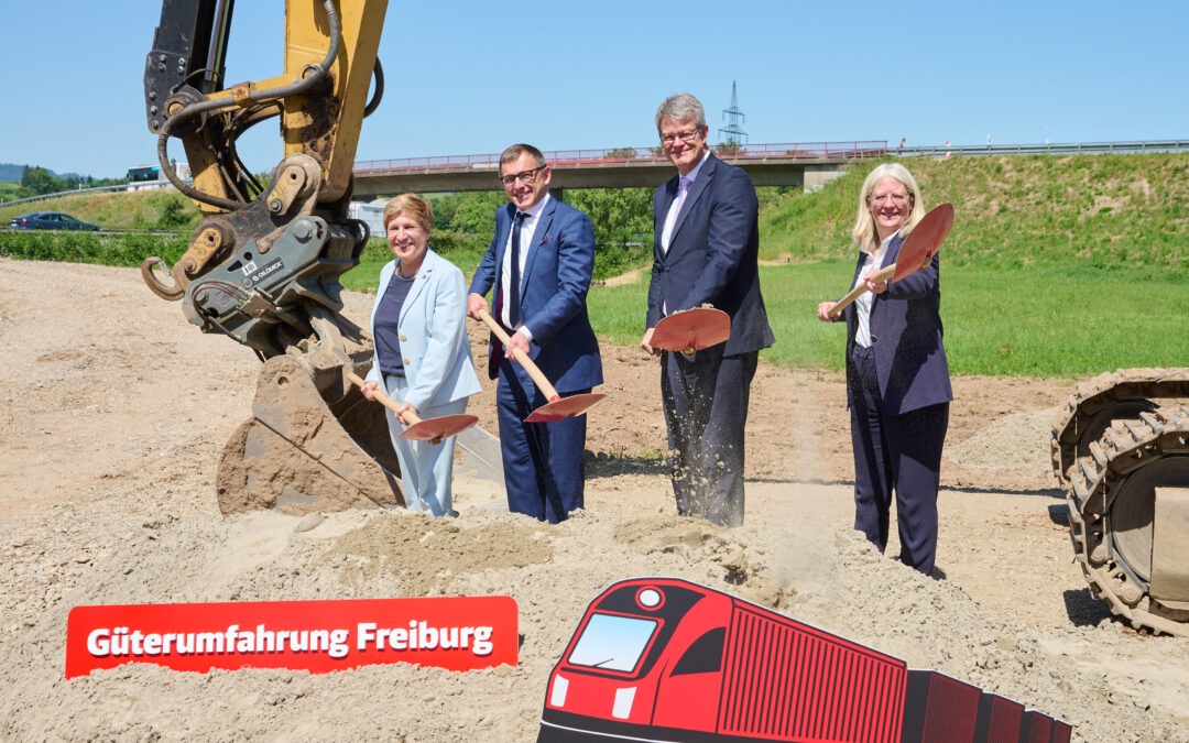 Four professionals in business attire with shovels stand at a construction site beside a large excavator under a blue sky, smiling for a photo.