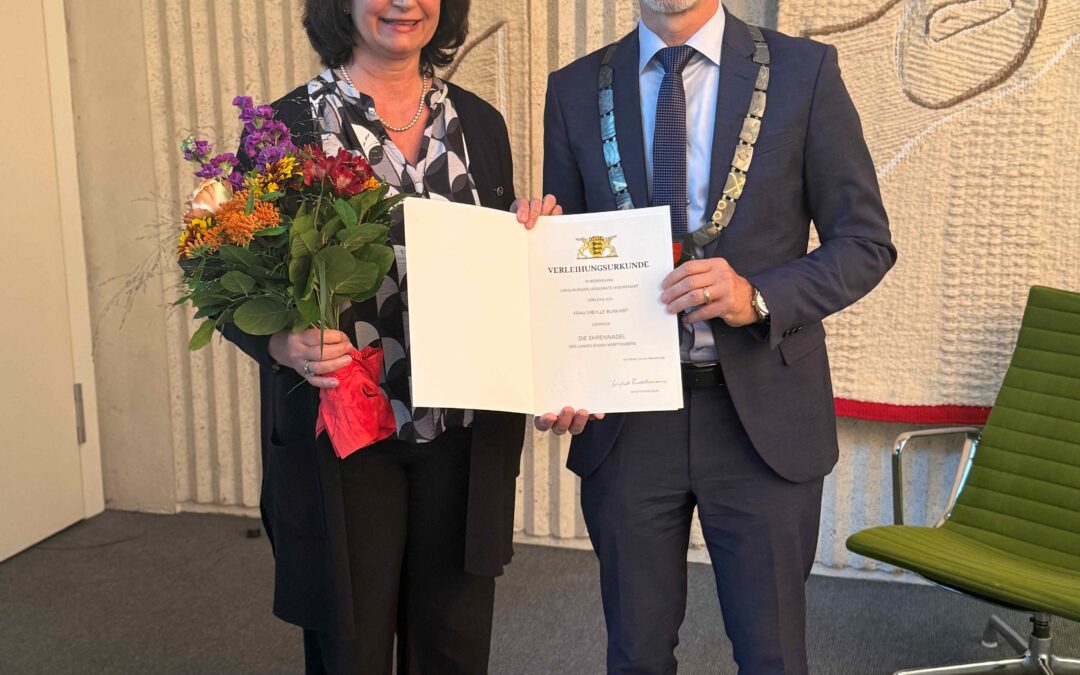 Two professionally dressed individuals posing with a certificate: a woman holding a bouquet of flowers and the document, a man in a suit wearing a ceremonial chain, both smiling indoors in front of a patterned wall.