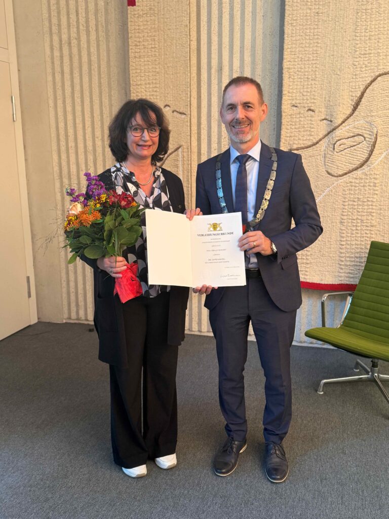Two professionally dressed individuals posing with a certificate: a woman holding a bouquet of flowers and the document, a man in a suit wearing a ceremonial chain, both smiling indoors in front of a patterned wall.