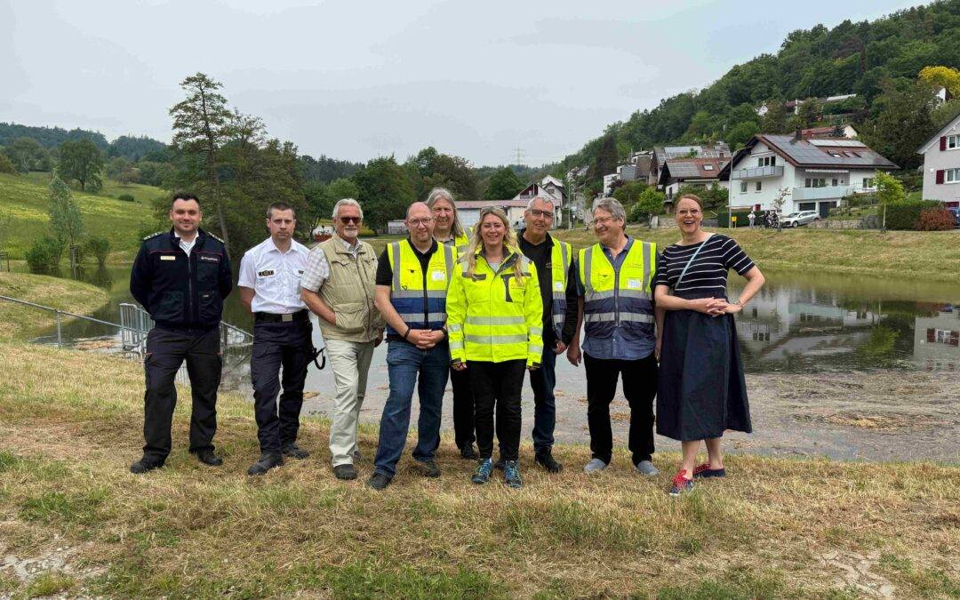 Group of eight people, some in high‑visibility vests, posing outdoors in a rural area with houses on a hillside and a pond nearby.