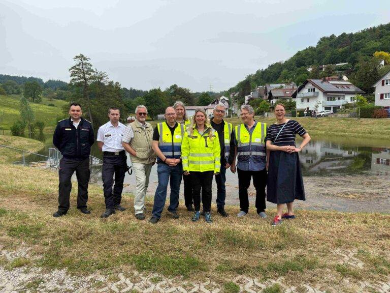 Group of eight people, some in high‑visibility vests, posing outdoors in a rural area with houses on a hillside and a pond nearby.